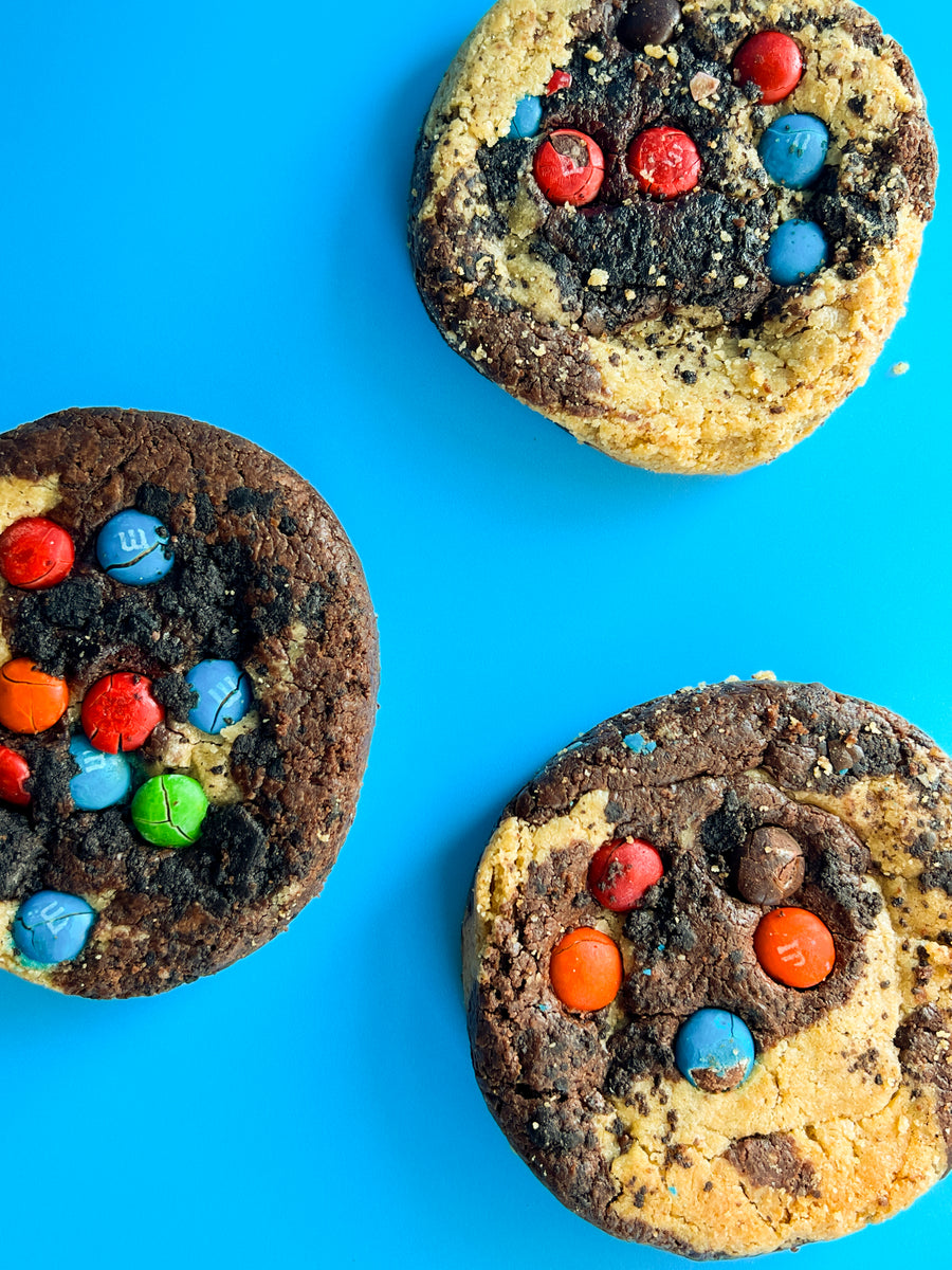 Three Brookie Monster cookies covered in candy pieces on a blue background
