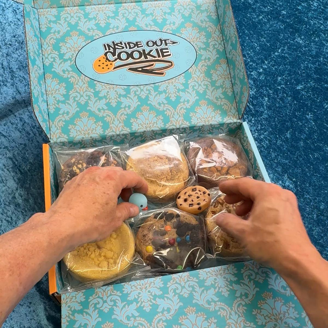 An open Inside Out Cookie box: damask lining, six cookies in sealed pouches, hands lifting a rubber duck